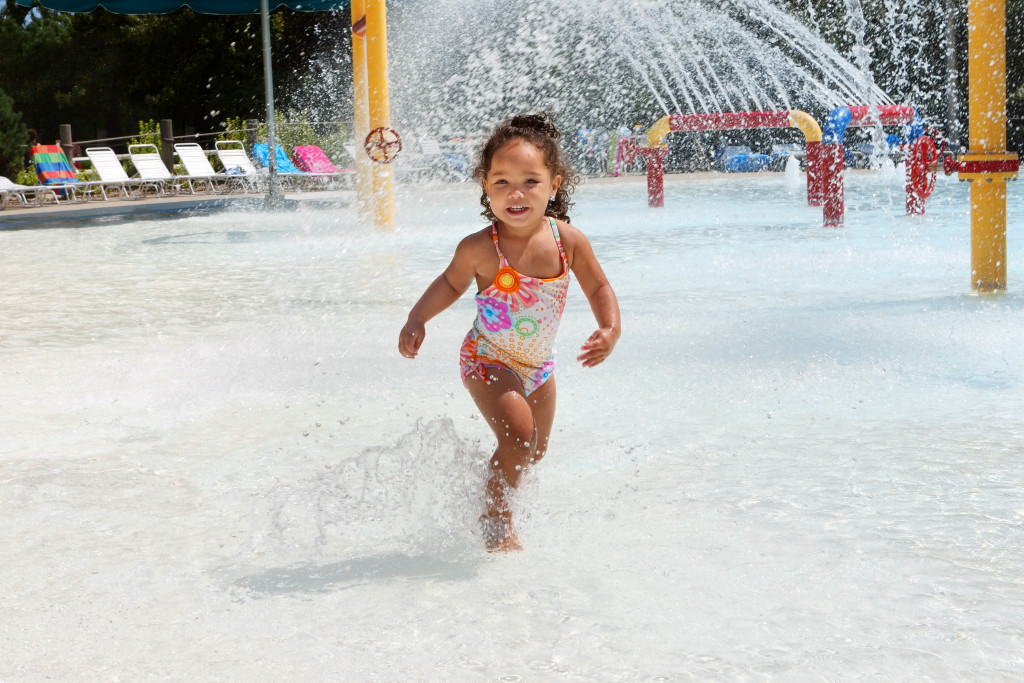 A child is standing in a pool and water is being sprayed in the background